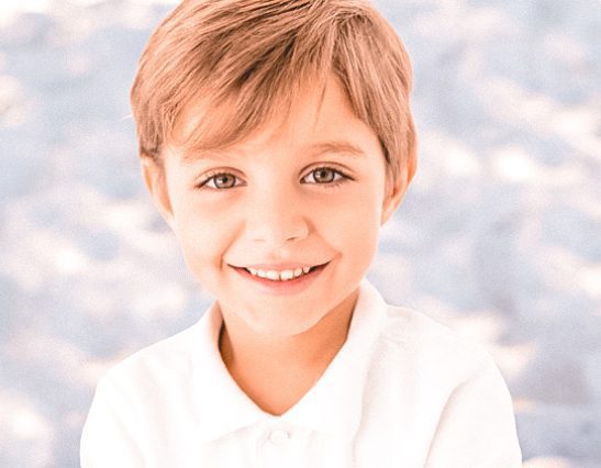 Little boy smiling with white collard shirt on and a blue background.