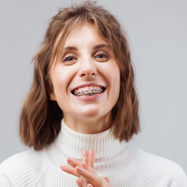 Woman in a white turtleneck smiling wearing metal braces. Light grey background.
