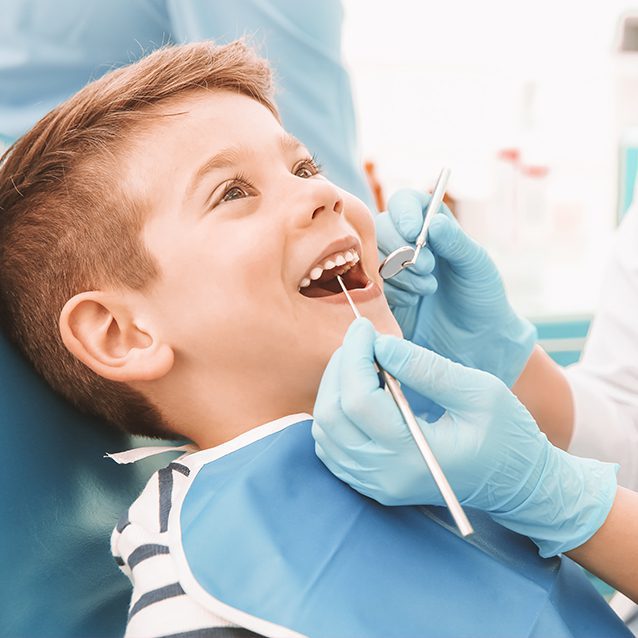 Young child in dentist chair with mirror and cleaning pick being held by dental assistant on blue background.