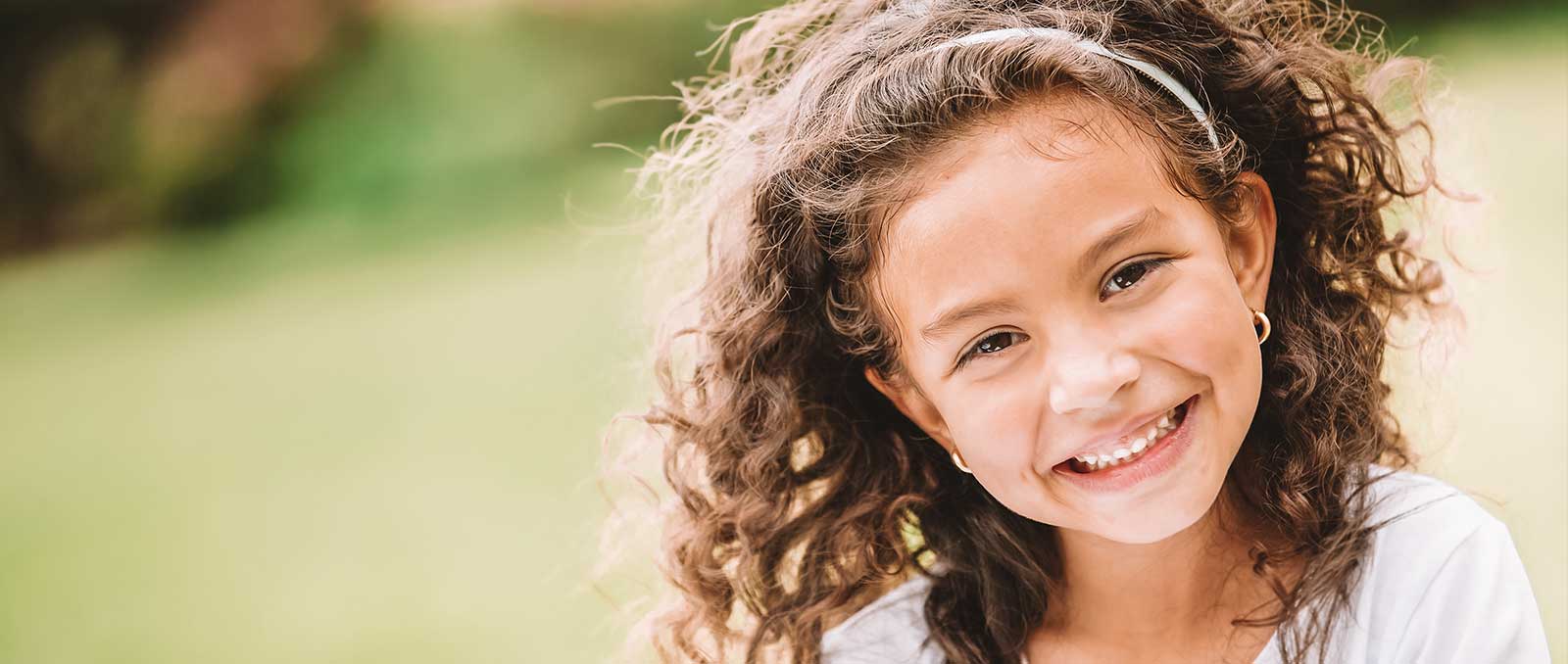 Little girl with curly hair smiling and sitting outside with a green blurred background.